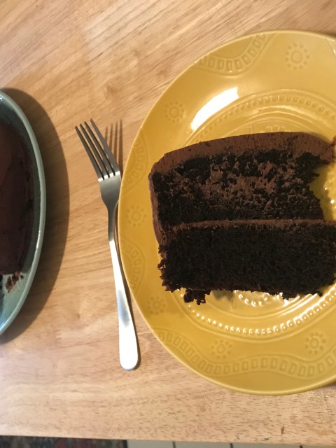 A homemade double chocolate layer cake. In the foreground is a slice of the cake on a yellow ceramic plate with a silver fork. In the background is the full cake (with the slice missing) on a light mint green plate. Both plates sit on a long wooden table.