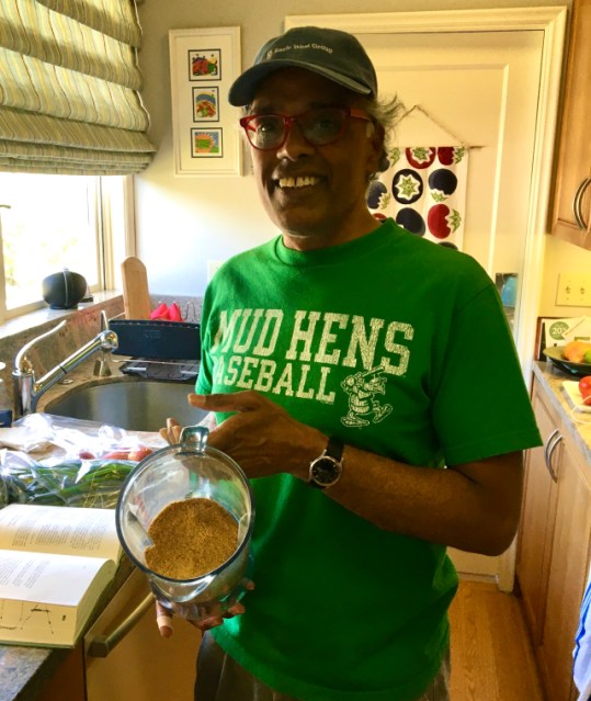 front shot of my dad, a south indian man in a baseball cap and bright green t-shirt that says Mud Hens baseball on it, holding up a finished batch of gunpowder in the blender