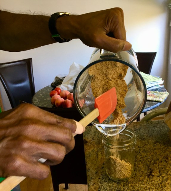 one brown hands shovels gunpowder from a blender into a small ball jar with an orange spatula