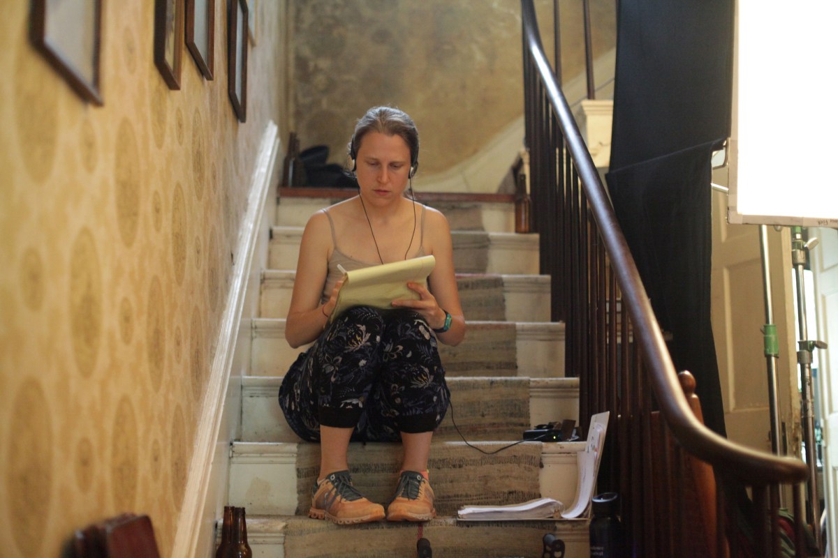 A candid photo of director Josephine Decker taking notes seated on a staircase on set