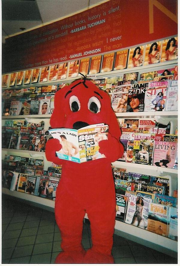 Stef dressed in a red Clifford costume reading a STAR magazine in front of a magazine rack in what looks like an old scanned photo taken with a digital camera