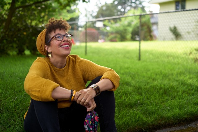 Kirsten Harris-Talley sits on the sidewalk with her knees up, one hand clasping the other, and gazes upward with an easy smile. She's sitting in  front of a green lawn that contrasts with the mustard yellow sweater and hat she's wearing.