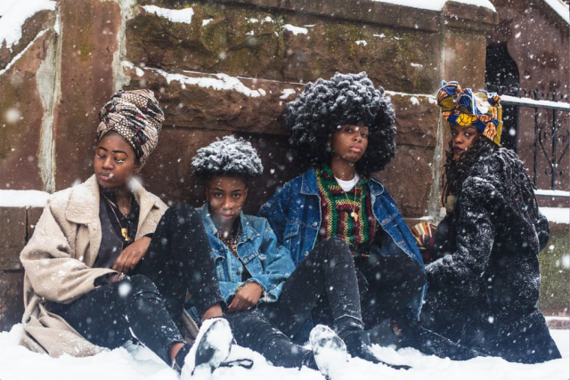 Four African Women sit in the wet snow against a brick wall. One woman has a brown headless and light brown wool coat. Two women have afros, covered in snow so they look more like crowns, and denim jackets. The last woman has a black hoodie wool coat and multi-colored head wrap.