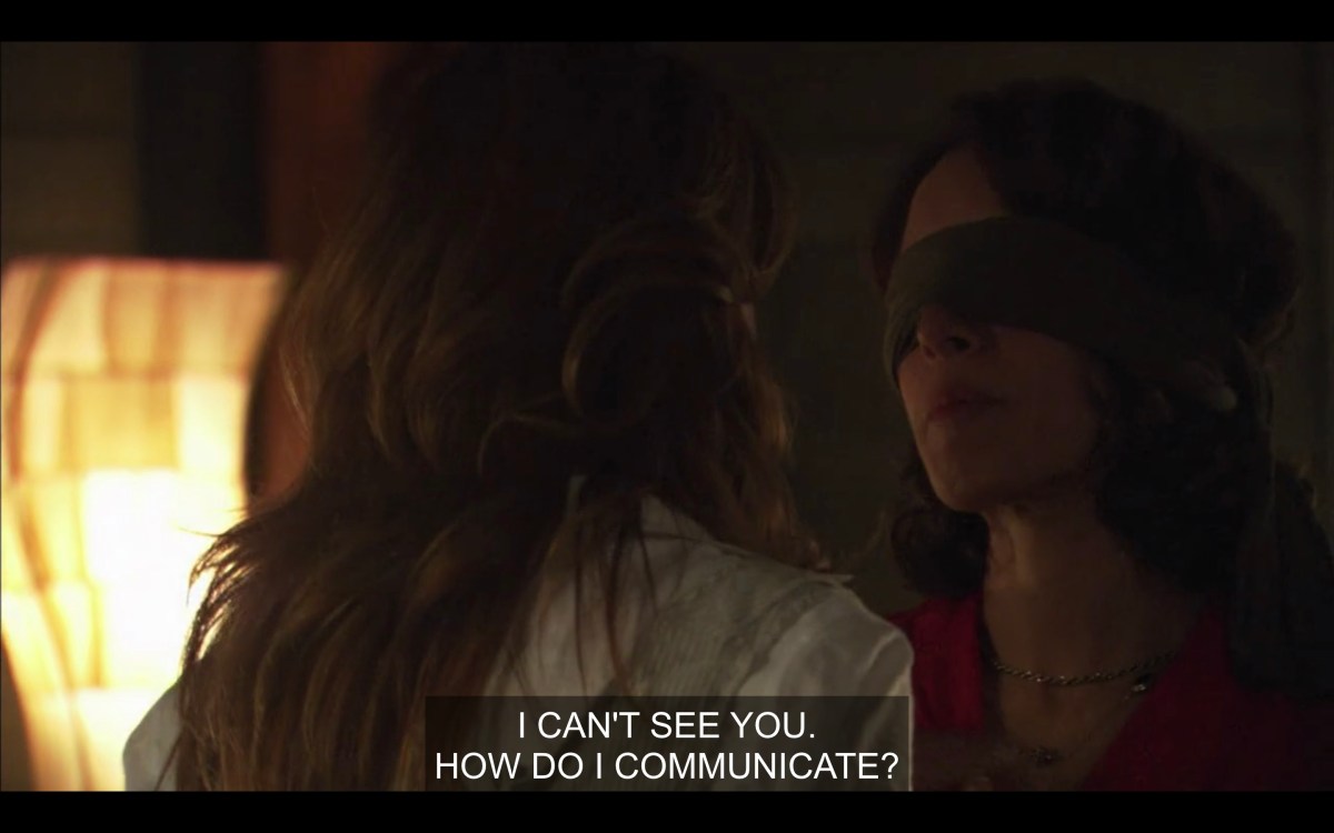 Jodi's back is to the camera, we can see Bette with a blindfold on. She is saying "I can't see you, how do I communicate"