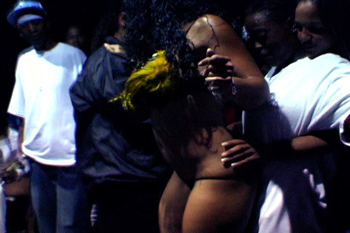 a Black woman in a g string and a sexy feathered top dances for a client in a white t-shirt, who is putting a hand on the dancing woman's waist