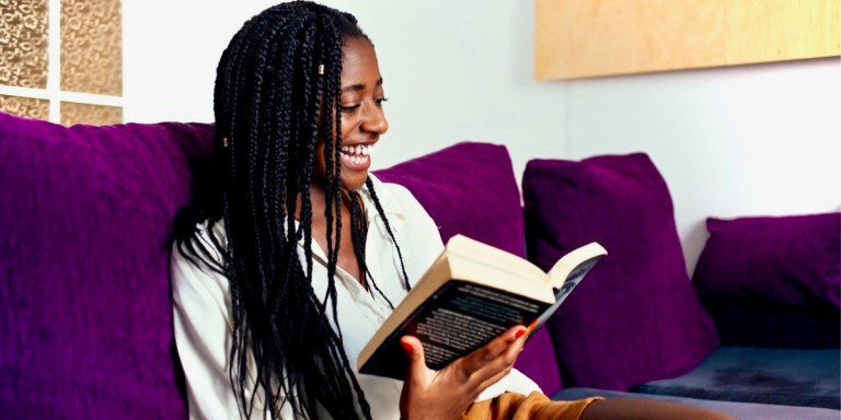 A cheerful young black woman with long braided hair is reading a thick book on a purple couch.