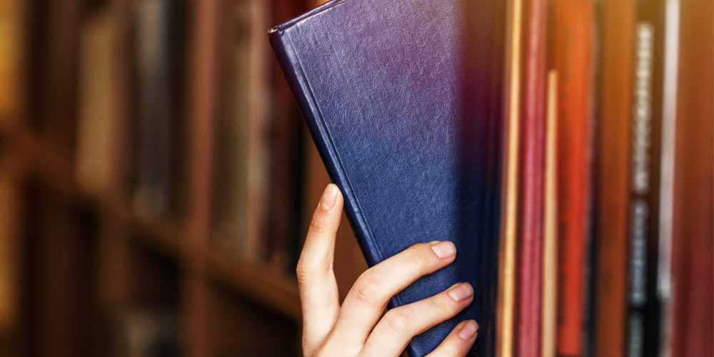 A photo of a light skinned person taking a library book off the shelf.