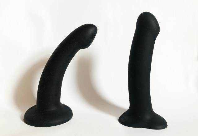 On the left, a curved, slightly tapering black dildo with a wide, solid circular base; on the right, a straighter, thicker black dildo with a straight shaft