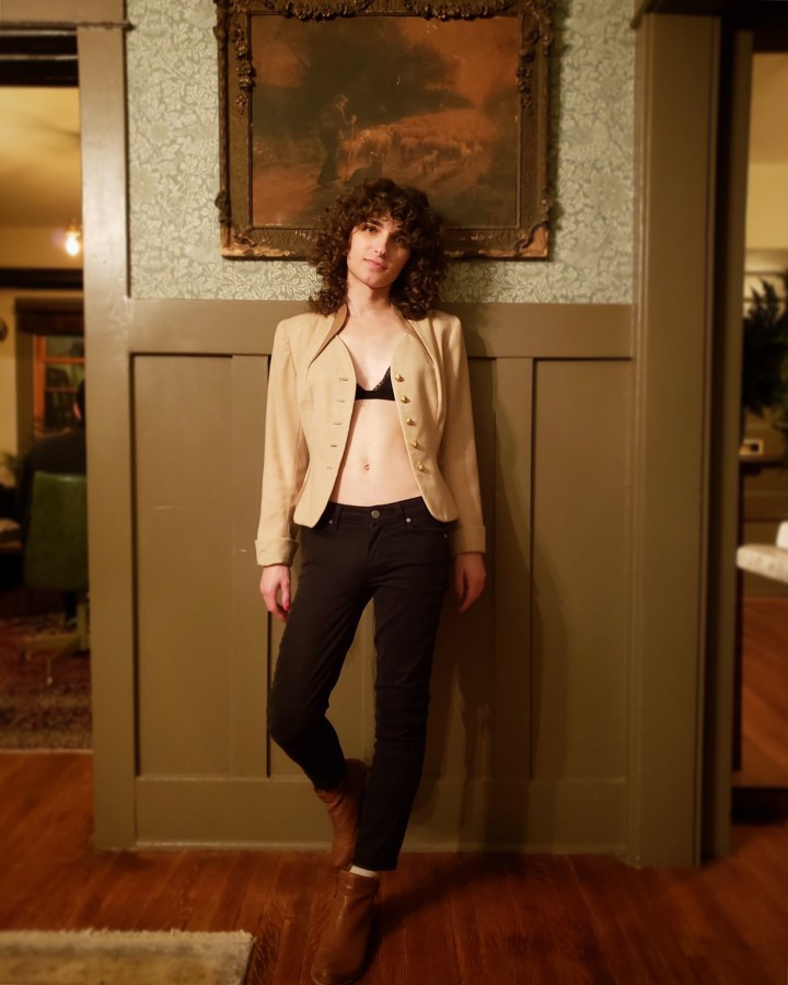 Drew is leaning up against a wall, with one foot propped up against the wall behind her; she's wearing black skinny jeans and ankle boots with a an open blazer worn over a black bra. 