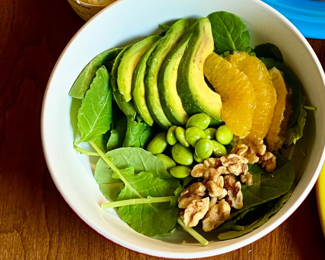 a shallow wide bowl with a bed of kale and fanned slices of avocado on top, slices of oranges nestled against those, a small pile of green edamame, and a small pile of walnuts 