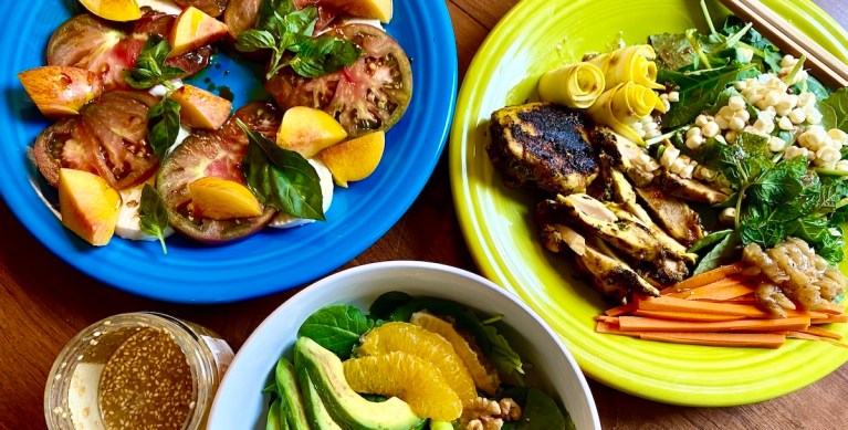 three dishes sit on a table touching their edges. a bowl with a peek of avocado and orange is at the bottom, a blue plate with tomatoes and nectarines is on the top right, and a bright green plate with chicken, corn, carrots and greens, and small rolls of yellow squash.