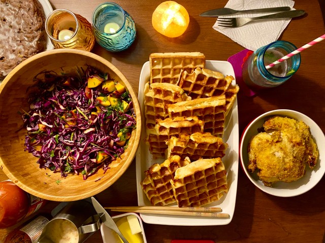 an overhead view of a table setting with a large bowl of purple cabbage salad, a tray of waffles, a pitcher of gravy, a small bowl with a little round fried hen nestled inside