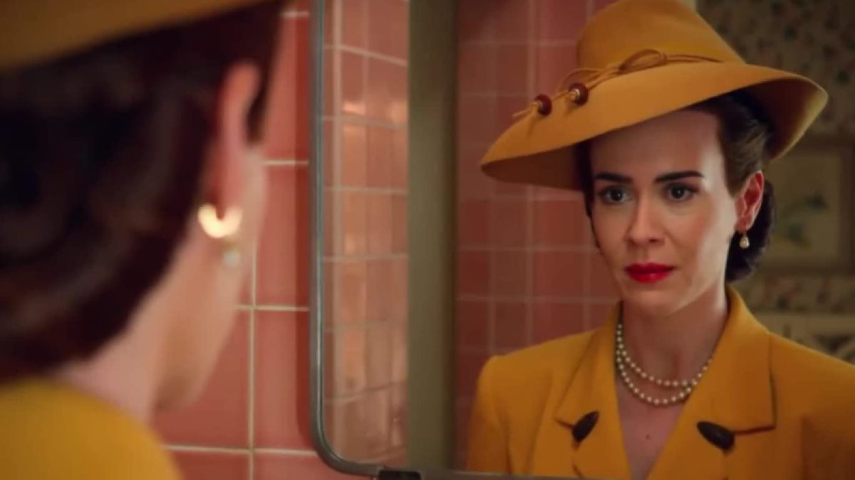 Sarah Paulson as Nurse Ratched in Ratched (showrunner Ryan Murphy, 2020). She is in a mustard yellow hat and dress styled like the 1940s and looks at herself in the mirror in a pink bathroom.