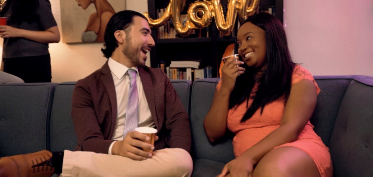 Two people sit on a navy blue couch in a party setting; on the left, a light-skinned man with longer hair and a short beard is laughing, wearing a formal jacket and a tie and holding a Solo cup. On the right, a Black woman with long straight hair and a light pink dress is making eye contact with him and laughing as she lifts a cup to her mouth.