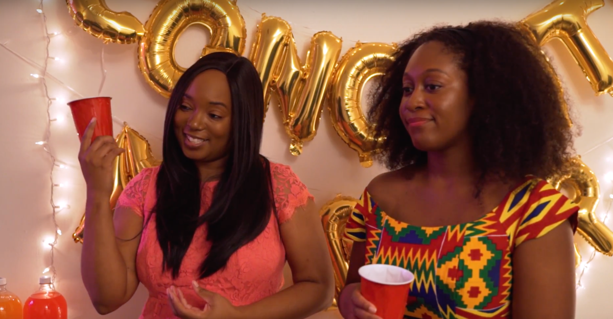 Two Black women stand next to each other smiling a bit stiffly in front of a golden balloon banner spelling CONGRATULATIONS; the woman on the left wears long straight hair and a bright colored top while gesturing with a red Solo cup; the woman on the right is wearing shoulder-length natural hair and a red, yellow and green patterned top while she holds her cup close to her chest, reserved.