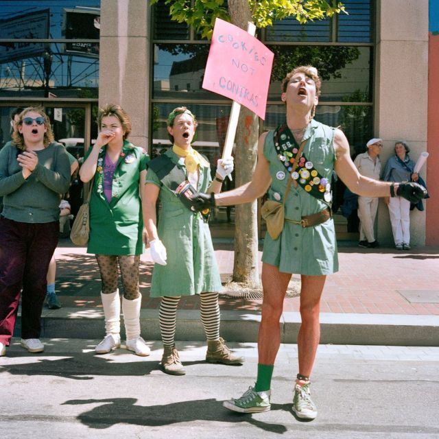 Adult queers wear Girl Scout uniforms in 1986 during a protest. In the background, one of the Girl Scout-clad protestors holds a sign that says "Cookies not Contras" in the trans pride colors.