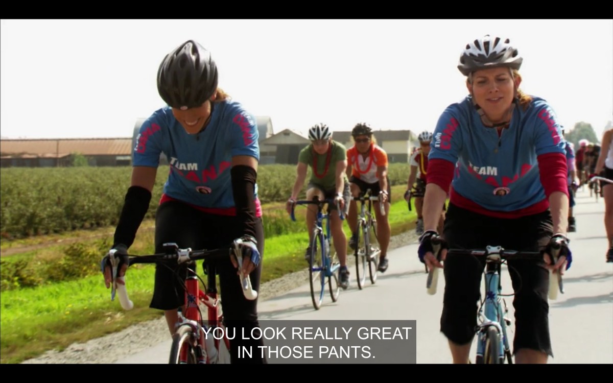 Bette and Tina biking, other bikers behind them, Pacific Northwest. Bette smiling, just told Tina her ass looks great in those pants