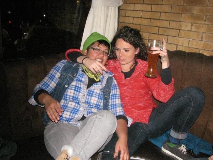 kamala and her friend amy sitting on a bench in a bar drinking beer. kamala is in an outdated flannel, demin vest and green beanie.