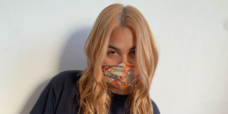 Hayley Kiyoko stands against a plain white wall. She has a pink multicolor mask and she's died her hair a new color, in the shade family of strawberry blonde.