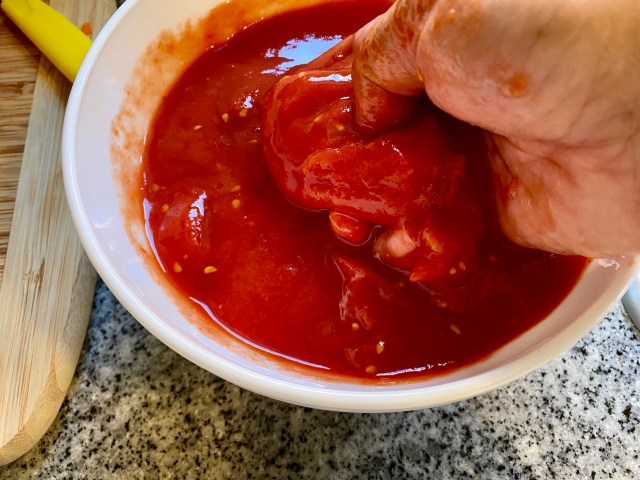 a hand half-submerged in a red bowl, squishing a tomato