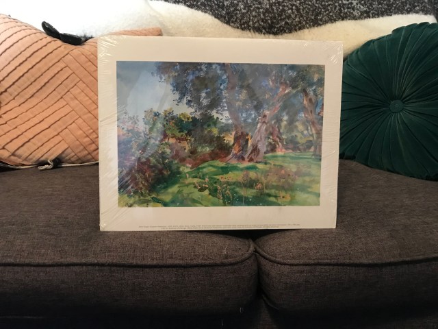 A brown couch with a blanket, a few throw pillows and an Impressionist landscape painting on it.