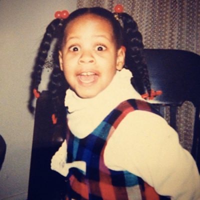 a young Black kid with braids and dressed in a plaid dress with a collar looks into the camera with wide open eyes and mouth partway open