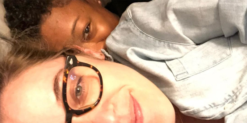 A close up selfie of Lauren Morelli laying in bed as the little spoon, with her wife Samira Wiley cuddling behind her as the big spoon. Samira's face is visible just behind Lauren's ear.