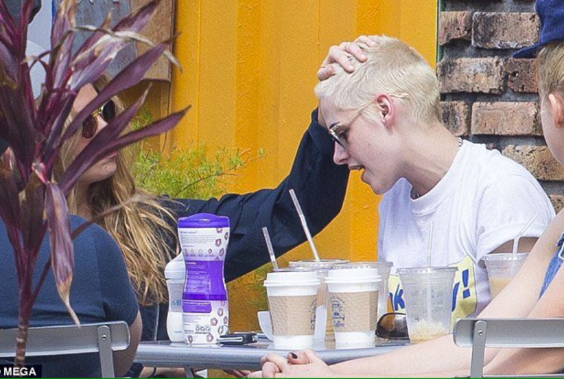 Seated at an outdoor dining table, Stella Maxwell rubs Kristen Stewart's buzzed blonde head.