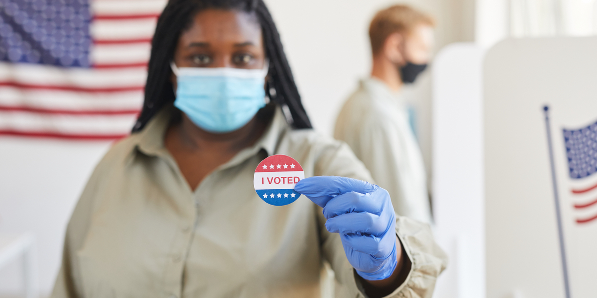 a black woman in a blue mask and gloves holds up a sticker that says