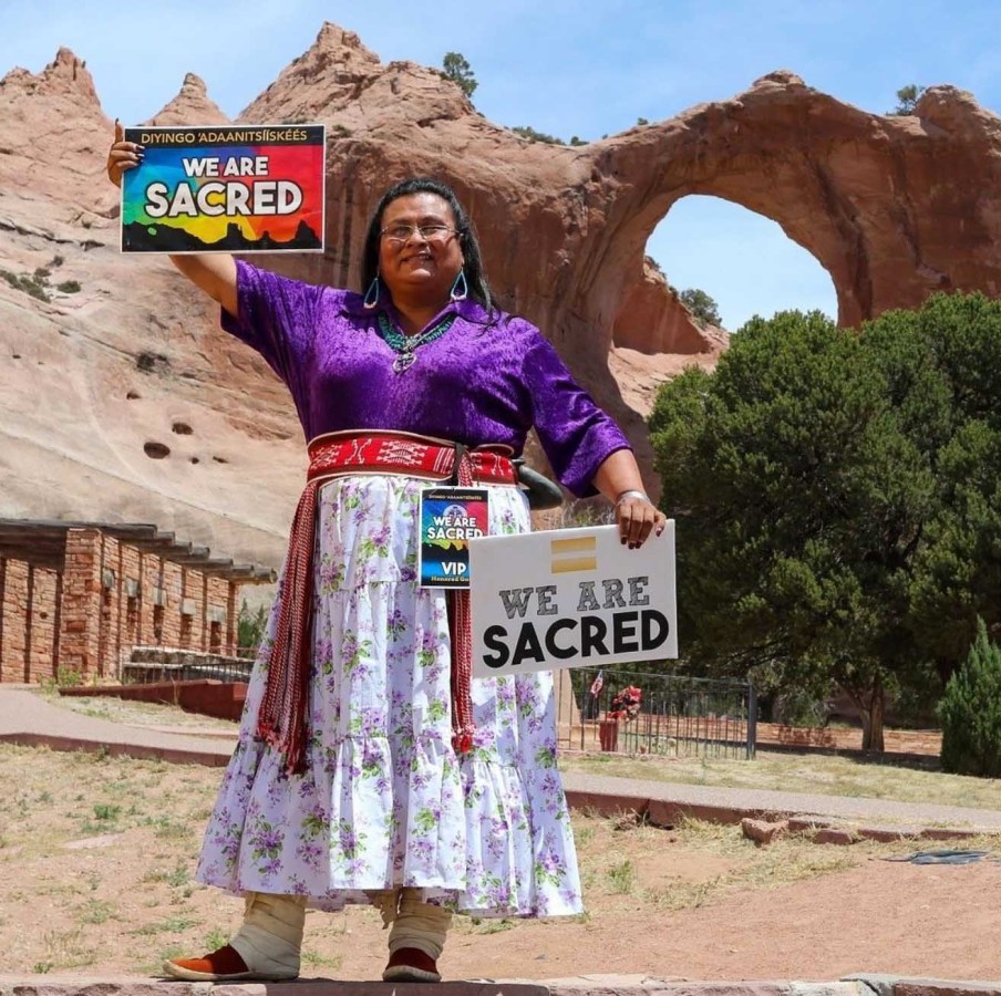 Mattee Jim stands in front of a canyon of rock, earth, and trees. She wears a rich purple shirt and a white skirt adorned with beaded belts. She holds two signs that read: "We are sacred."