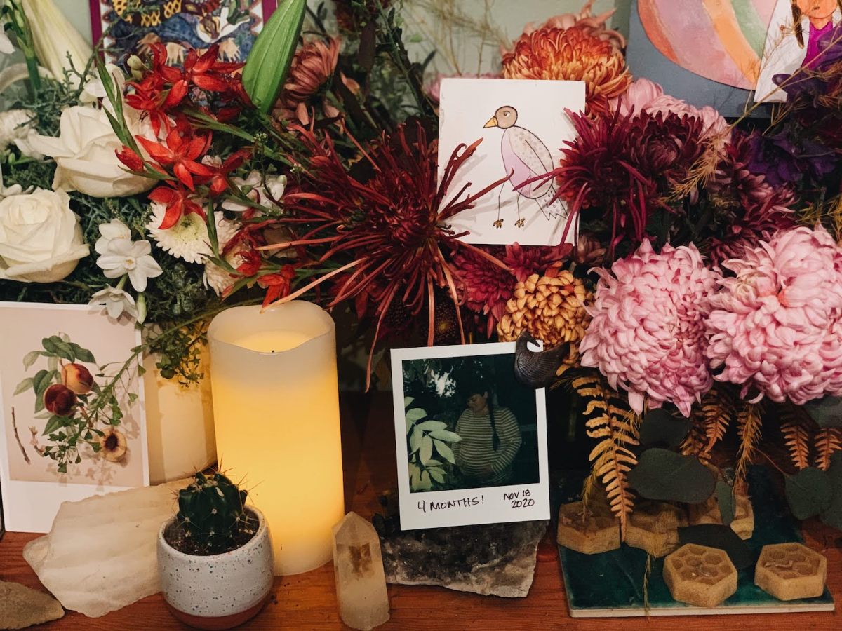 a grief altar filled with flowers, a candle, a drawing of a tiny bird, and a polaroid of a pregnant woman captioned 4 months!