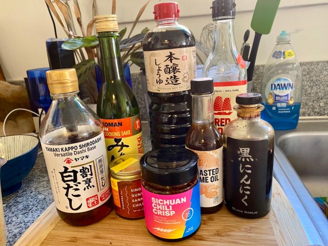 a line up of sauces in jars and bottles: dashi, mirin, chili powder, chili crisp, soy sauce, fish sauce, sesame oil, garlic vinegar