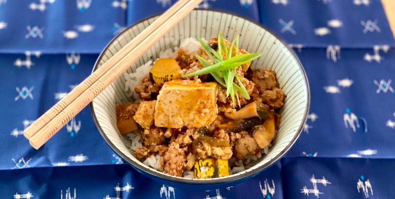 a bowl of mapo tofu over rice topped with a star of sliced scallions. chopsticks rest on the left side of the bowl and the bowl in on a blue woven cloth.