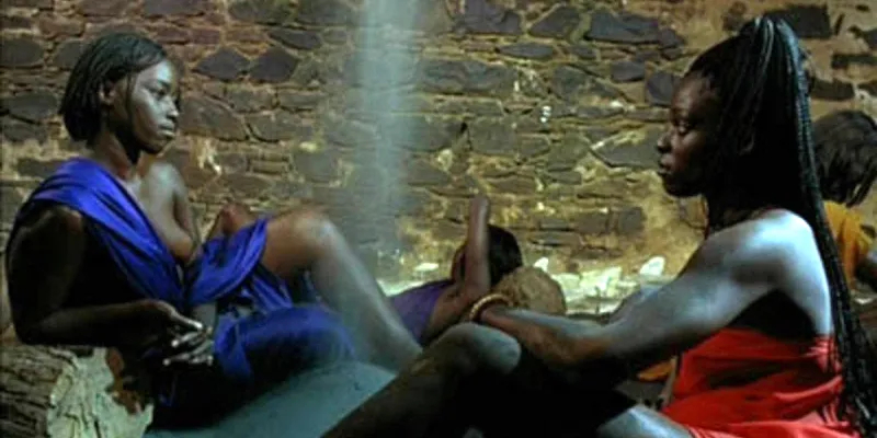 Two Black women, one in a bright blue wrap and the other in a bright red wrap, look at each other against a stone backdrop.