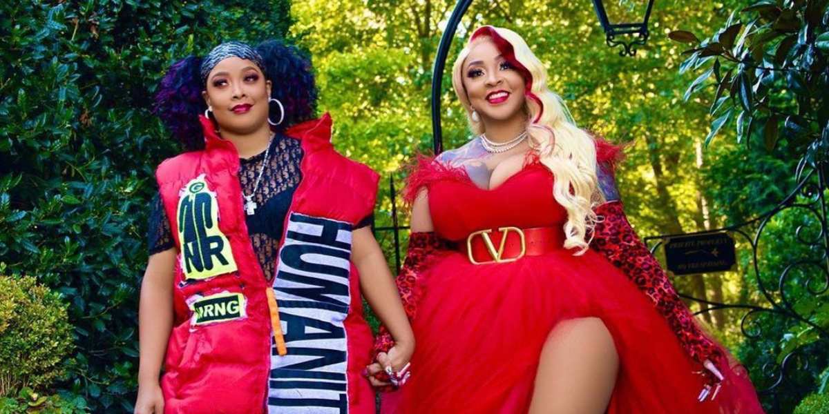 The rapper Da Brat in a bright puffy vest, and her wife is holding her hands in a off shoulder red dress with a high slight on her thigh. They look happy and in love, with green trees glowing alit in the background.