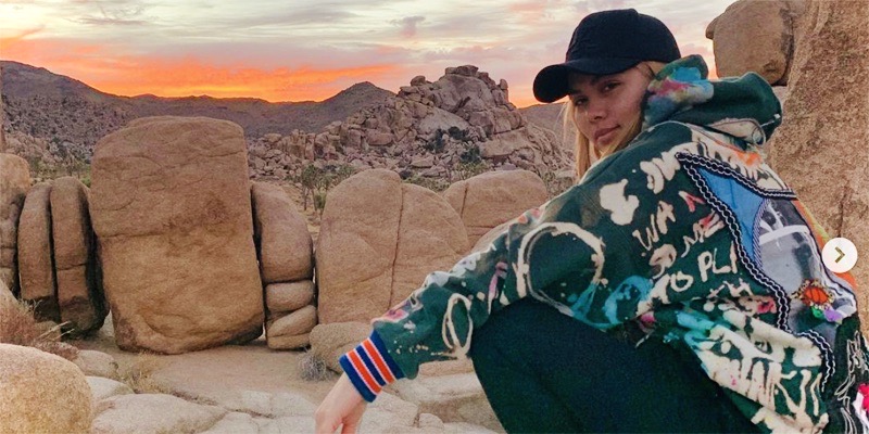 Hayley Kiyoko in a color jacket and a baseball cap squats against sun kissed boulders and mountains as the sun sets in orange behind her.
