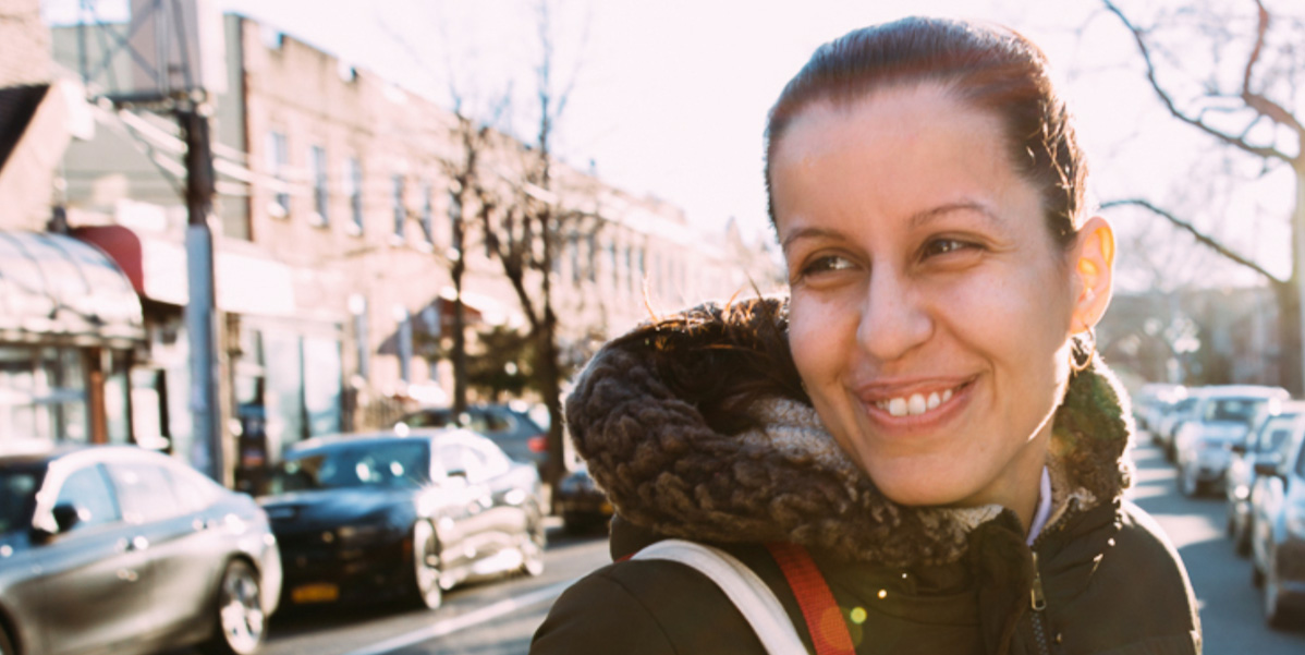 A photo of Tiffany Cabán, a queer latina with a long pony-tail, looks over her shoulder and her parka hood as she crosses the street.