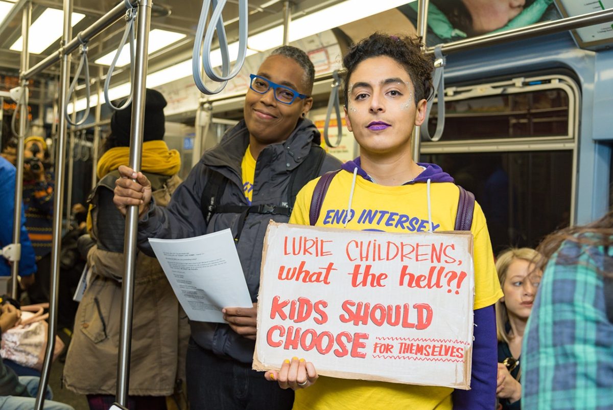 Sean Saifa Wall (left) and Pidgeon Pagonis taking over a public train to protest against intersex surgeries at Lurie Children's Hospital in 2018. Sean Saifa has dark brown skin, short curly hair, and wears glasses. Pidgeon has light skin, wavy hair, and has on purple lipstick.