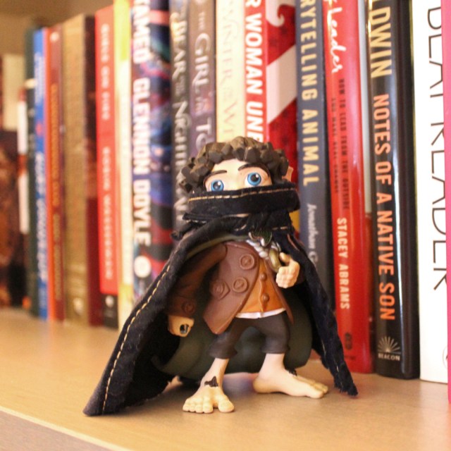 A small toy Frodo Baggins is standing on a bookshelf wearing a mask and a cap. Frodo is the Anti-Fascist on the Shelf!