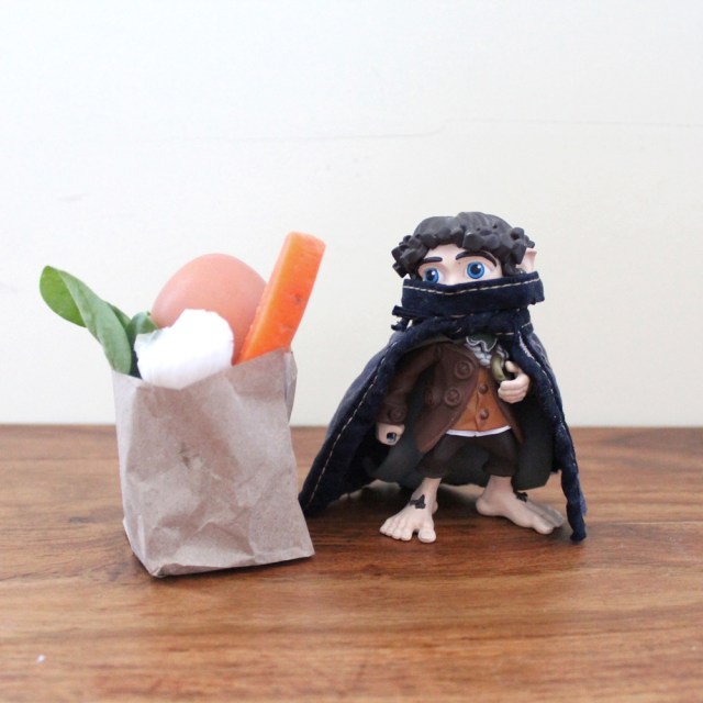 A small toy Anti-Fascist in a face mask and cap stands triumphantly next to a paper bag filled with fresh produce.