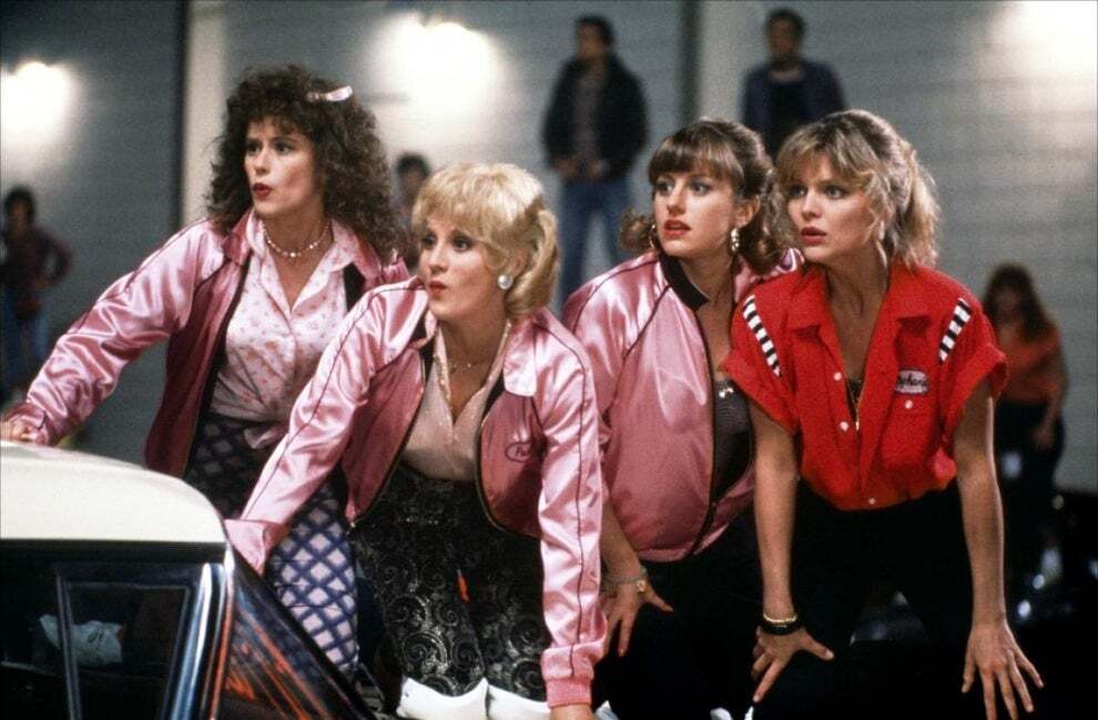 Still from Grease 2. 4 girls wearing Pink Satin Jackets and one in a red bowling shirt, all lean atop a car.