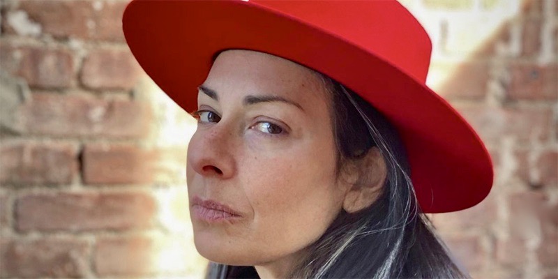Stacy London in a boss looking bright red wide brimmed hat against a brick background with just a perfect streak of sunlight against her face.