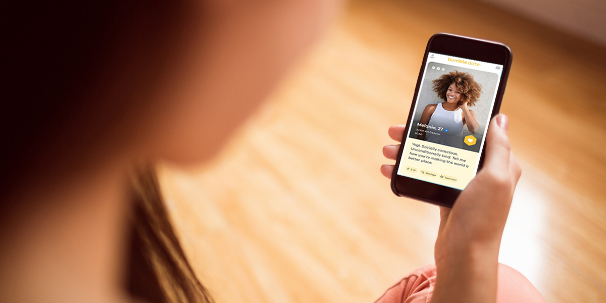 A woman sitting on a hardwood floor browses Bumble on her phone.