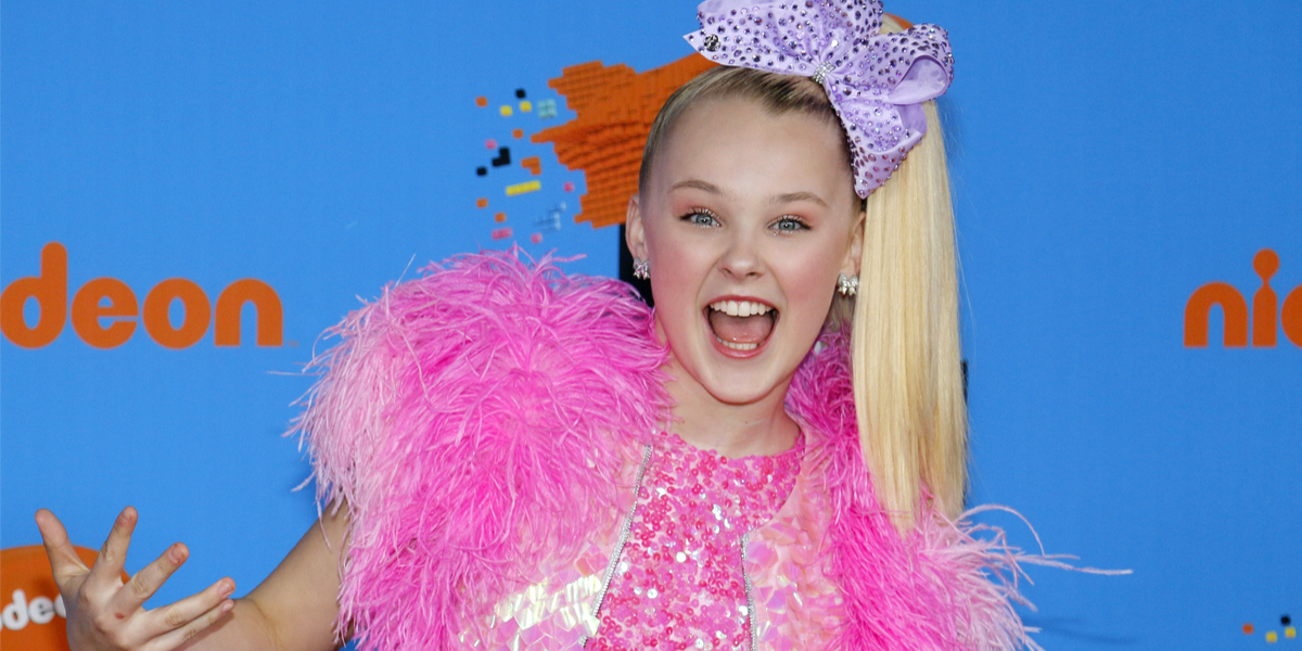 JoJo Siwa wears a purple hair bow and pink outfit made of feathers and sequins.