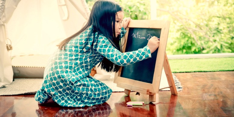 A young girl is in a green dress and bent off a chalkboard as she works on a math problem against a spring background, it looks serene.