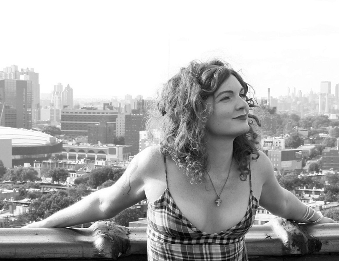 A black and white photo of Bryn Kelly, a women with culry hair wearing a cross necklace and sundress, smiling on the roof of a building