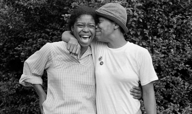 Two black lesbians from the 1970s hold each other while laughing and smiling for the camera in this black and white photo. One of them is in a button down shirt with rolled up sleeves and their hair in a bun. The other one is in a white tee and a hat.