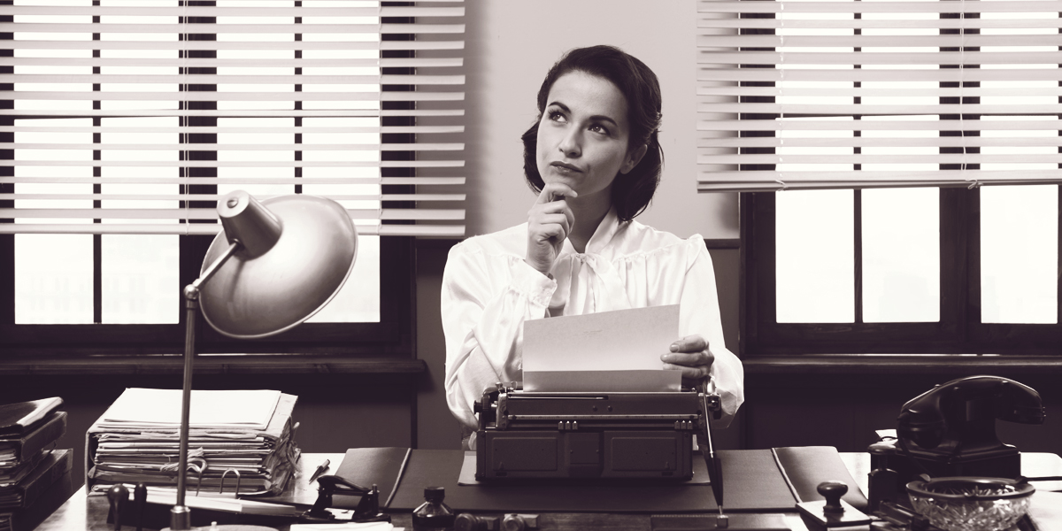 A woman sits at a typewriter and holding her chin pensively.