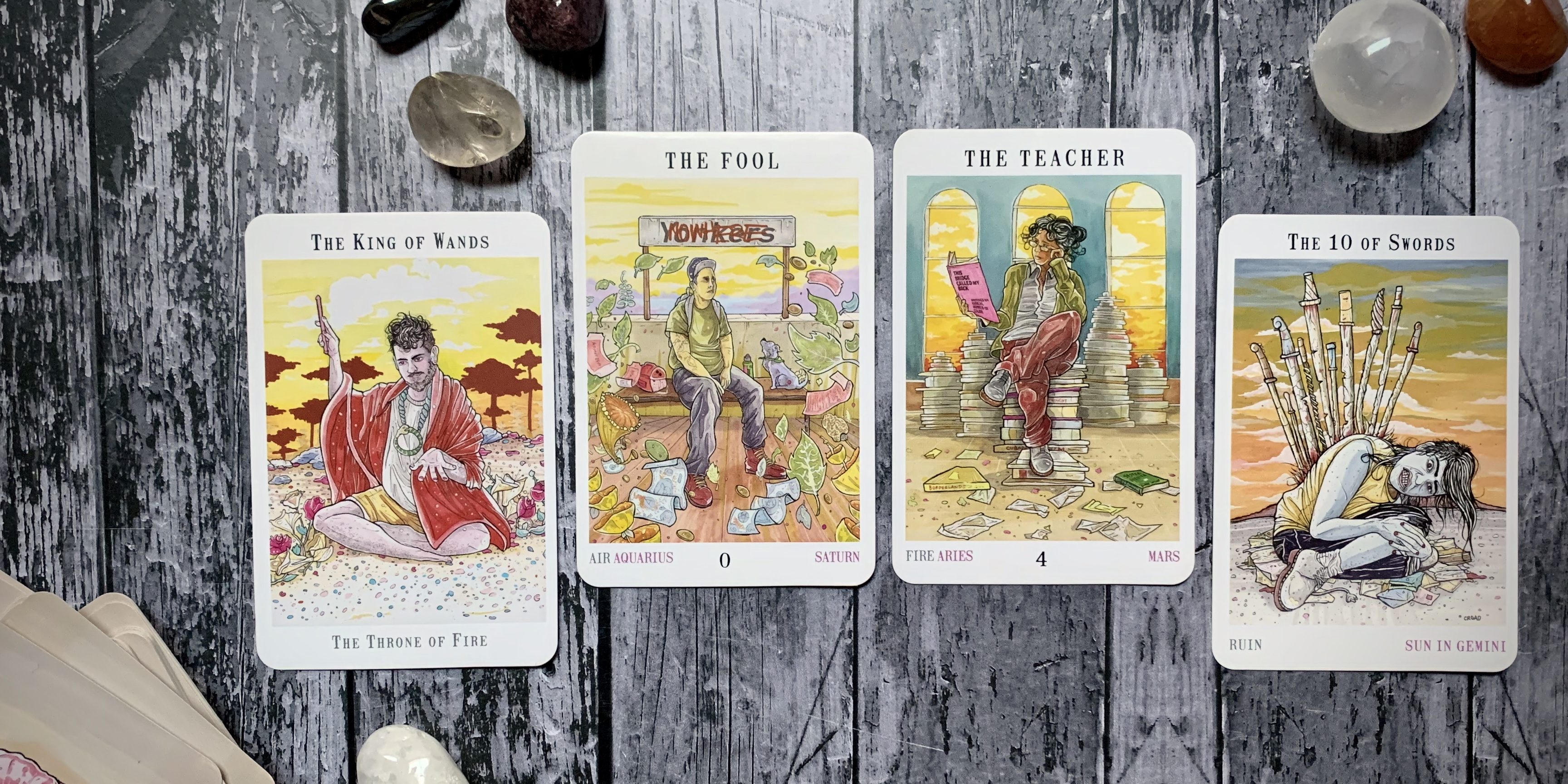 Four tarot cards from the Next World deck arranged on a wooden surface surrounded by some stones and crystals; from left to right, the cards are the King of Wands, the Fool, the Teacher and the Ten of Swords.