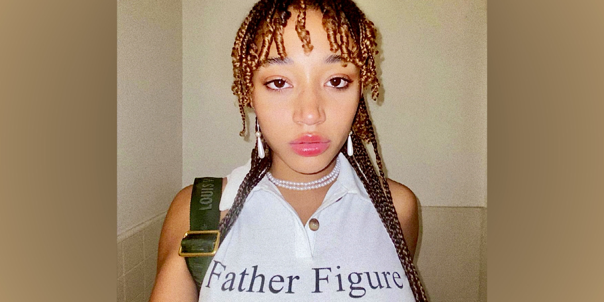 Actor Amandla Stenberg is in a white tank top that has "Father Figure" written in black letters. They have pink lip gloss and curly hair tendrils around their face.